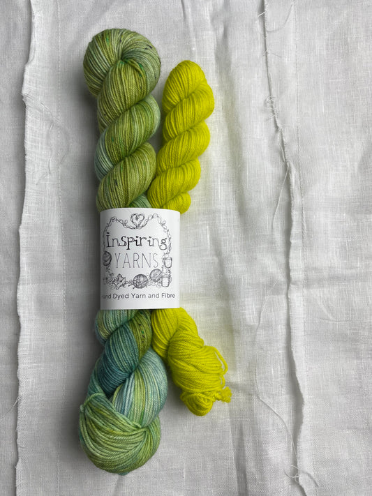 A set of hand dyed sock yarn comprising one 50g variegated light blue, green and teal and one 20g bright acid yellow on a white linen background.  The yarn is branded Inspiring Yarns.