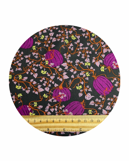 Round fabric swatch of Pumpkin Patch Deep, from the Spooky 'n' Witchy Collection by Art Gallery Fabric, a pattern of purple pumpkins and vines on a dark background with a wooden ruler in centimetres for scale, on a white background.