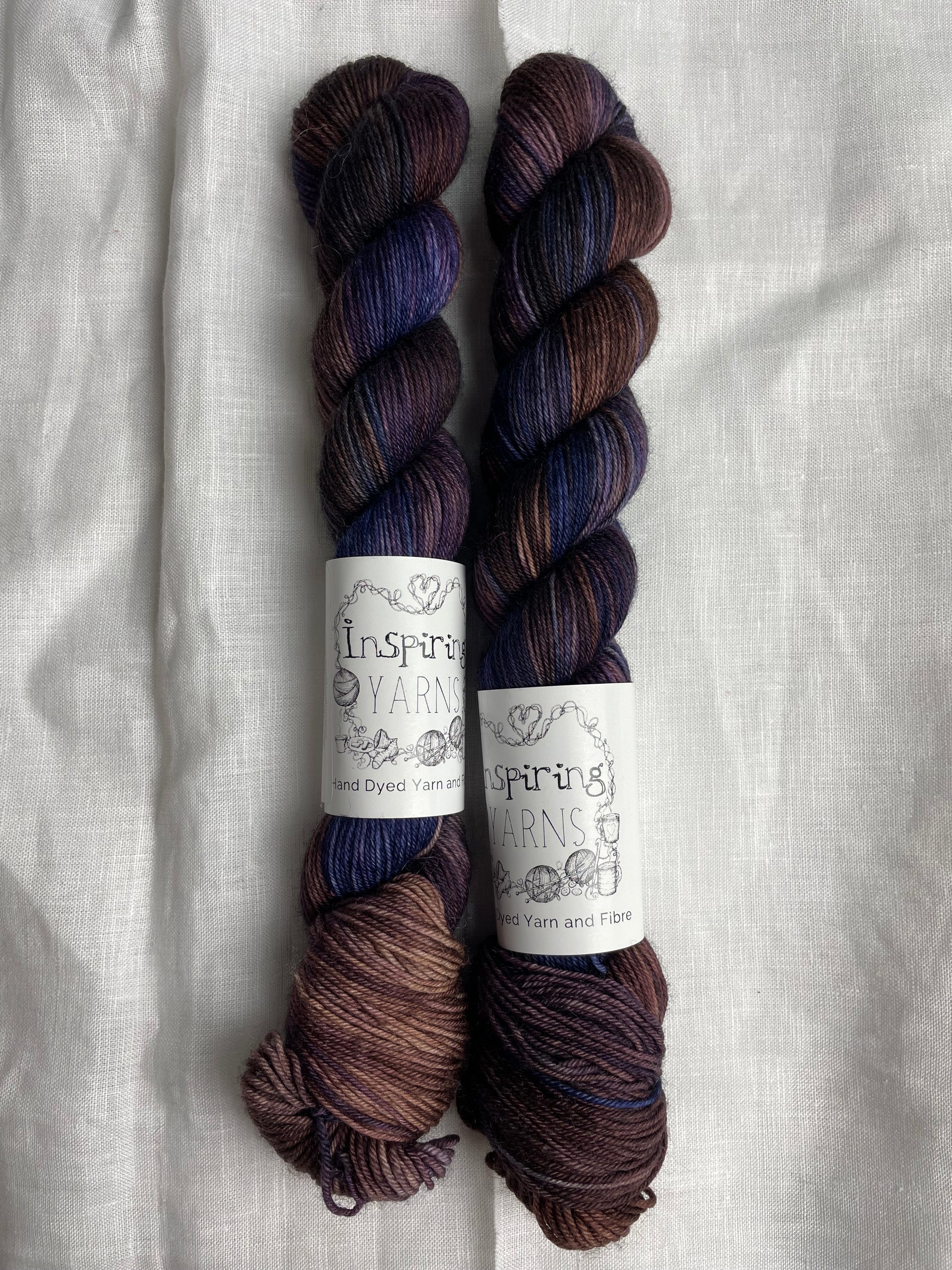 Two skeins of Denise, a navy, brown and purple coloured Merino Smooth Sock, labelled Inspiring Yarns on a white linen background. 