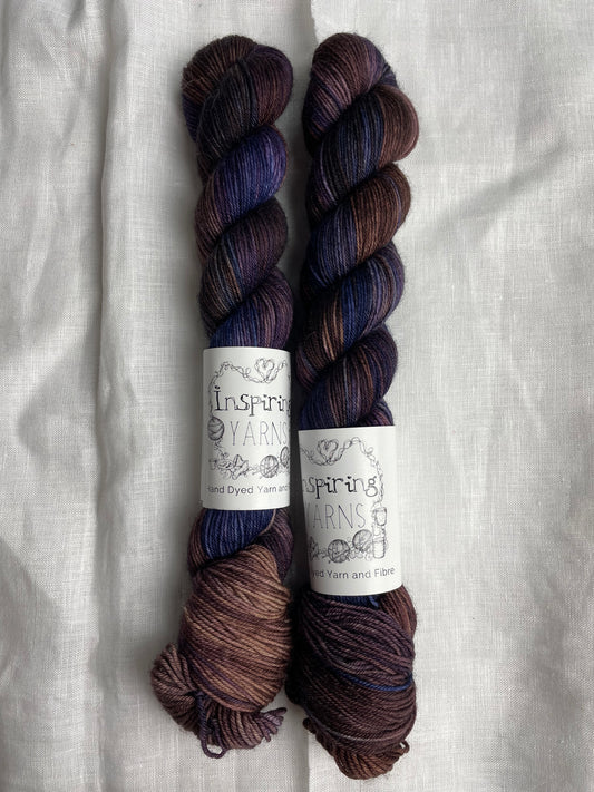 Two skeins of Denise, a navy, brown and purple coloured Merino Smooth Sock, labelled Inspiring Yarns on a white linen background. 