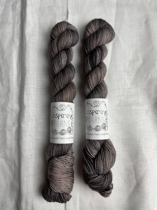 Two skeins of Merino Smooth Sock yarn hand dyed in a tonal grey colourway and labelled Inspiring Yarns on a white linen background.