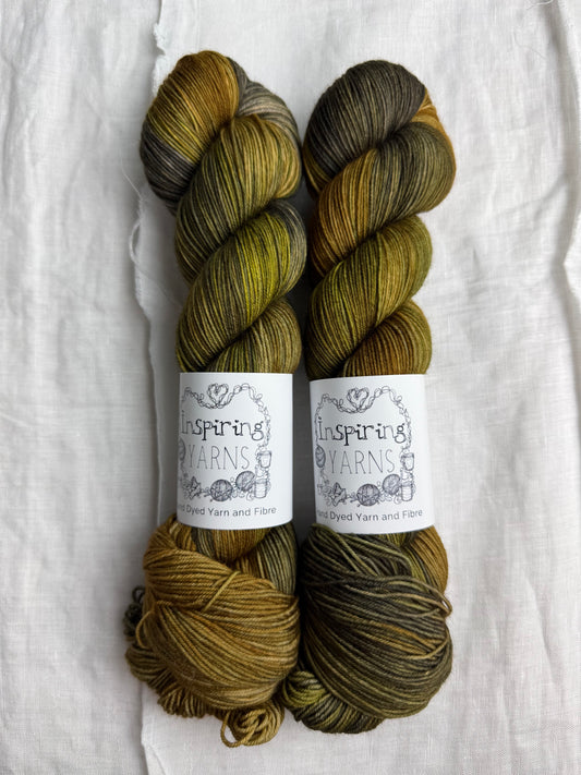 Two skeins of variegated dark grey, green, yellow and brown yarn with Inspiring Yarns labels on a light textured background.