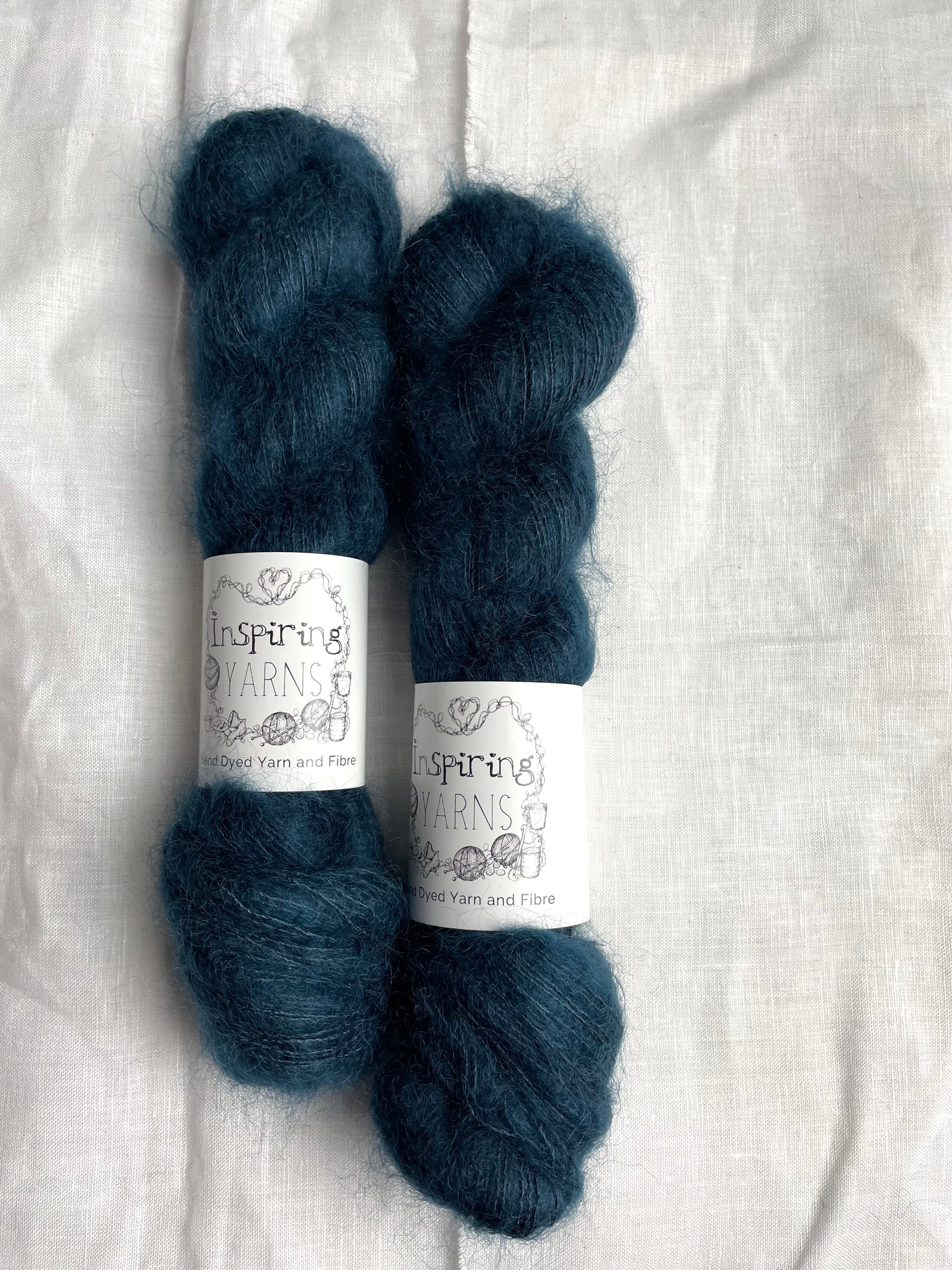 Two skeins of dark steel blue mohair silk lace yarn labelled Inspiring Yarns on a white linen background.