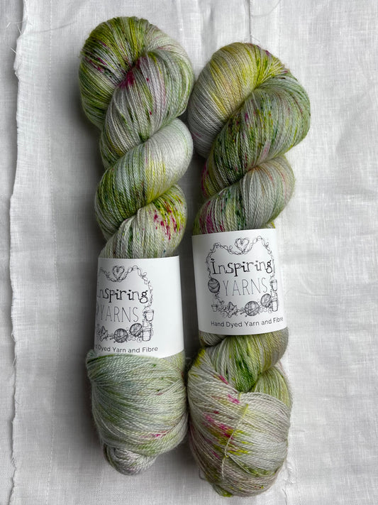 Two skeins of pink and green speckled silver grey coloured hand dyed Merino Lace yarn branded Inspiring Yarns on a white linen cloth.