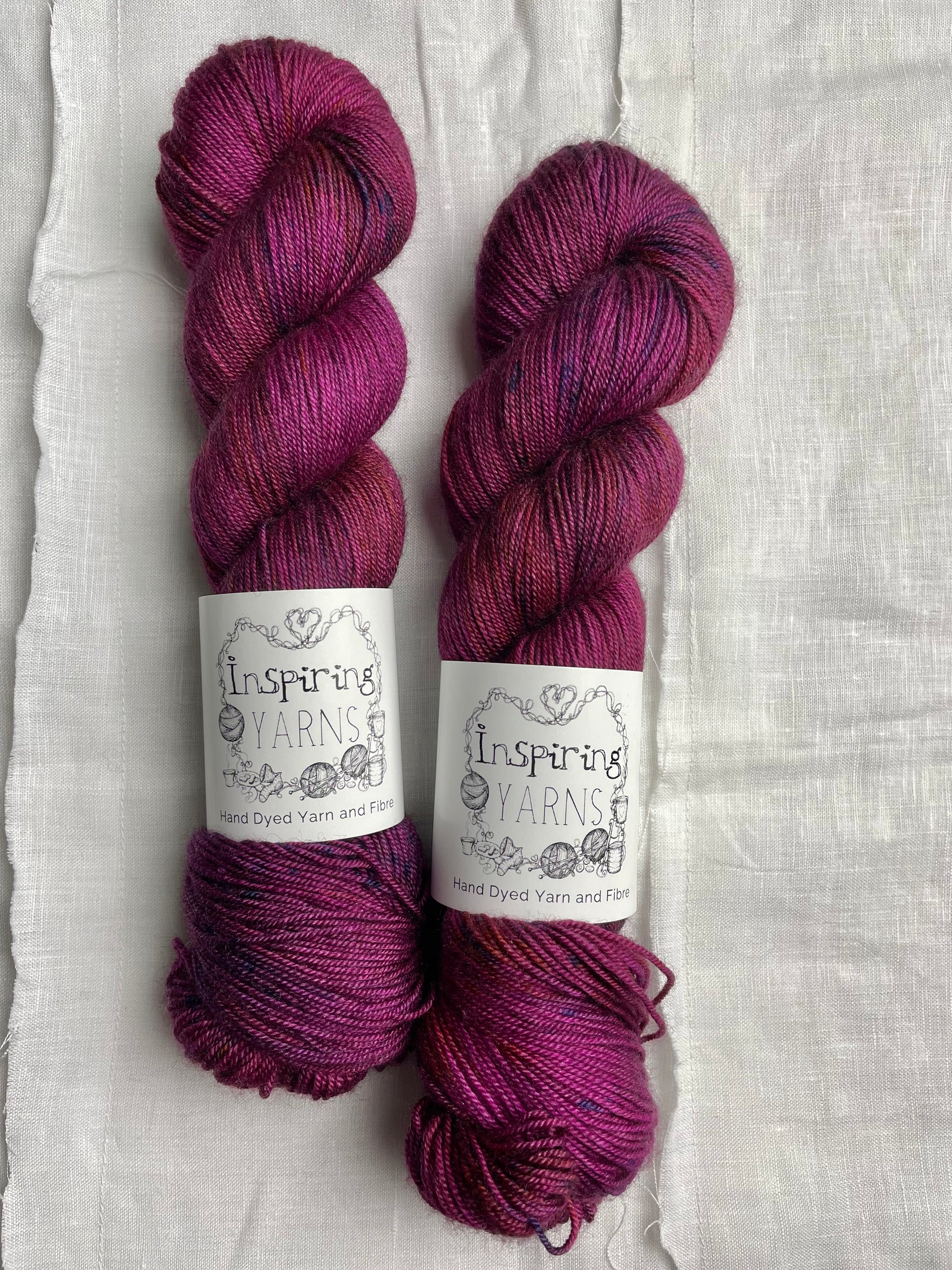 Two speckled fuchsia pink coloured skeins of hand dyed Yak Silk 4Ply yarn branded Inspiring Yarns on a white linen background.