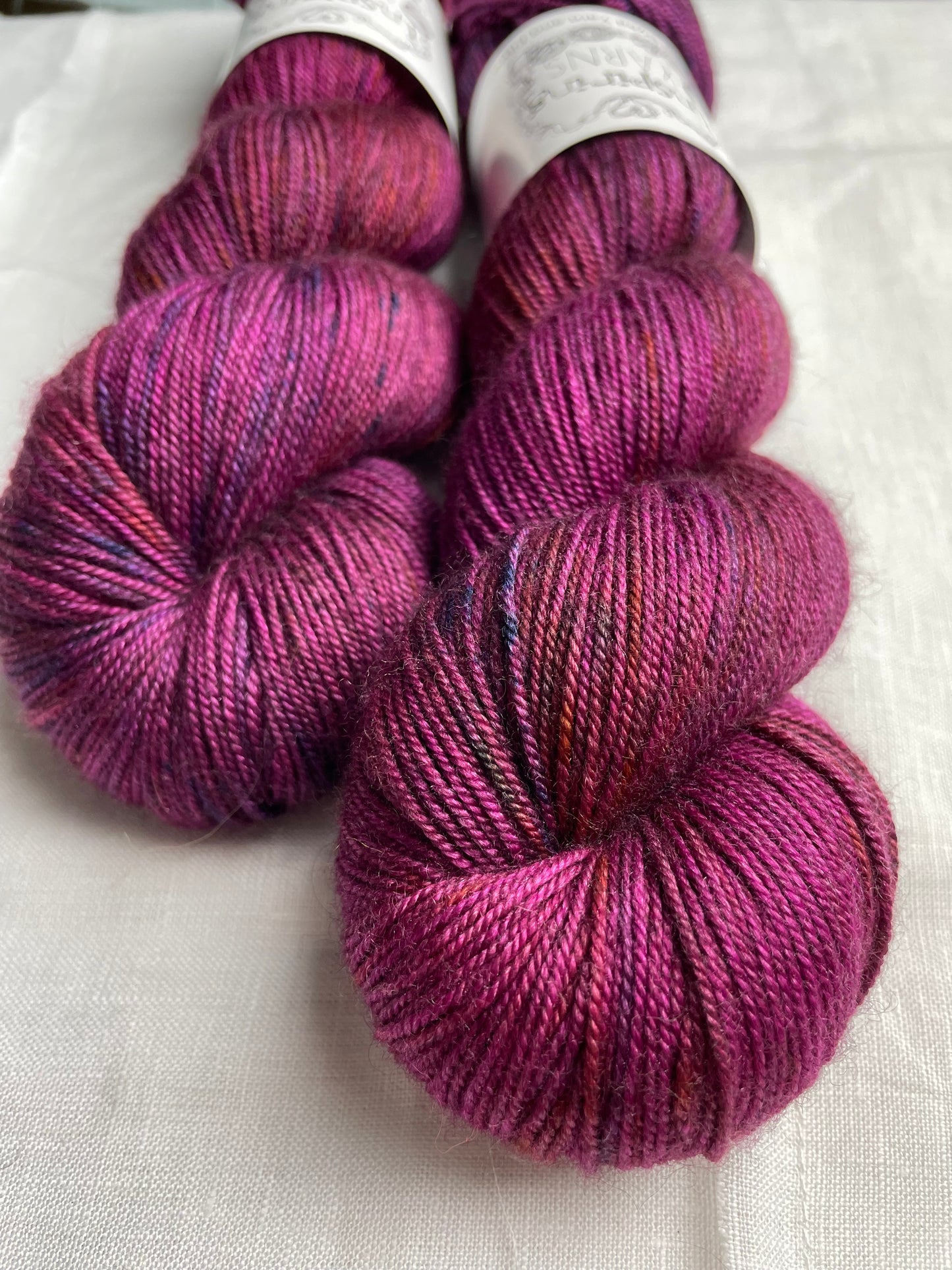 Close up of two speckled fuchsia pink coloured skeins of hand dyed Yak Silk 4Ply yarn branded Inspiring Yarns on a white linen background.