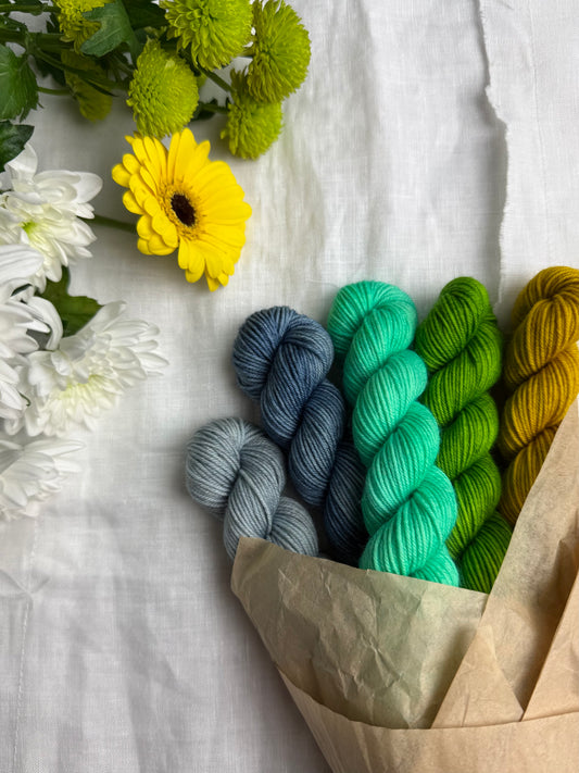 Five mini skeins of yarn in blue, blue grey, yellow, green & bright mint green, wrapped in brown paper, on a white background with flowers