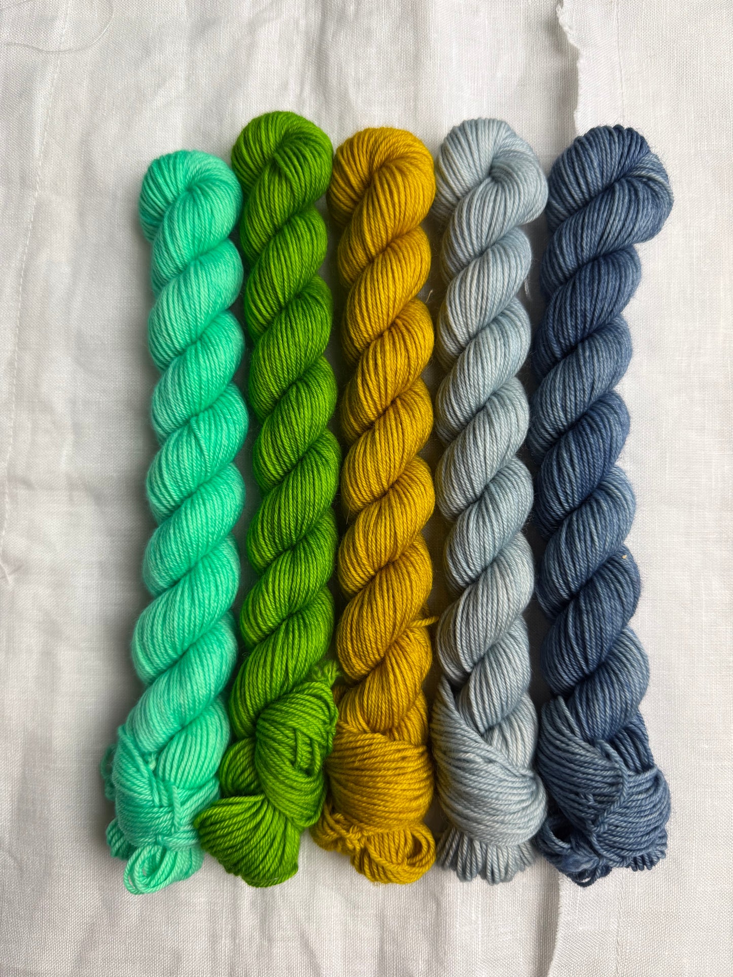 Five mini skeins of yarn in blue, blue grey, yellow, green & bright mint green on a light background