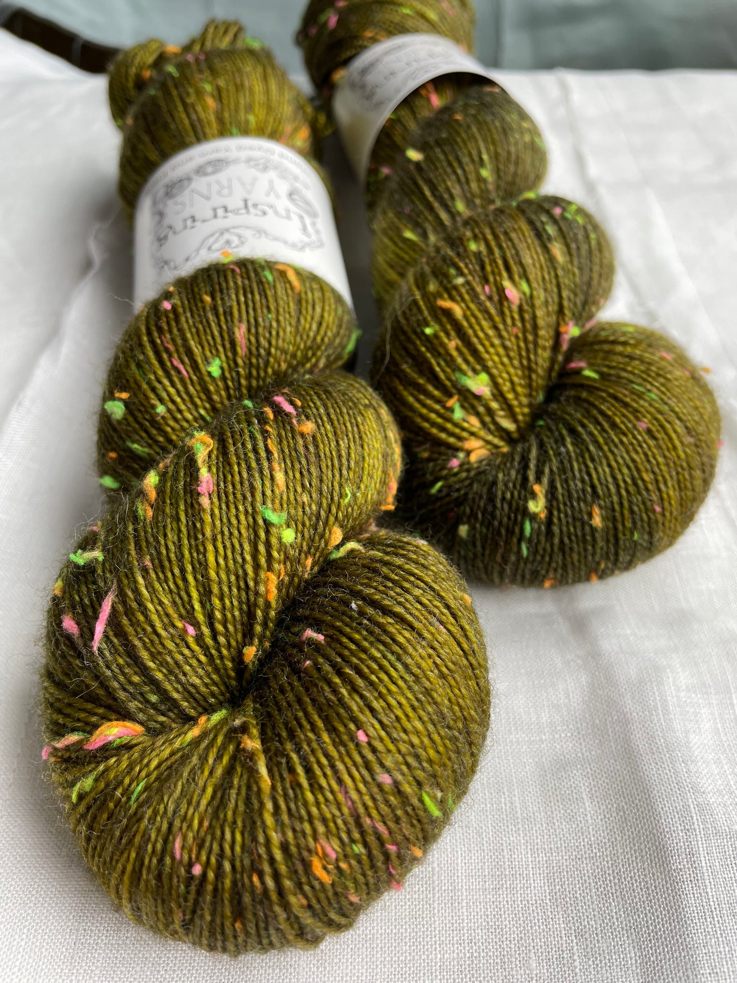 Close up of two skeins of tonal dark olive green colour  hand dyed Disco Donegal yarn branded Inspiring Yarns on a white linen background.  The yarn has multi coloured neon neps.