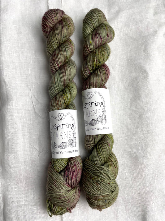 Two skeins of Cassidy, a sage green with speckles coloured Merino Smooth Sock, labelled Inspiring Yarns on a white linen background. 