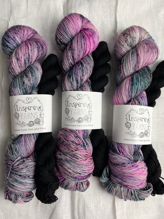 Three sock sets made of a skein of pink black and teal speckled hand dyed yarn with a contrasting black mini skein, labelled Inspiring Yarns and laid on a white linen cloth.