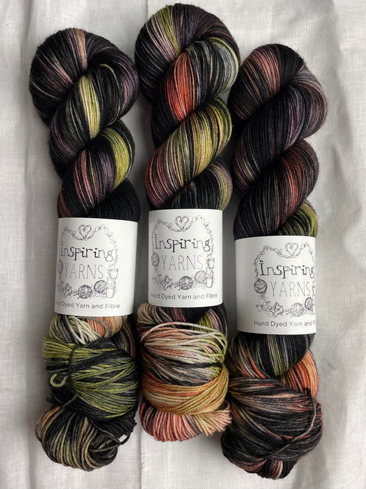 three skeins of black with lilac green and orange hand dyed yarn, labelled Inspiring Yarns and laid on a white linen cloth.