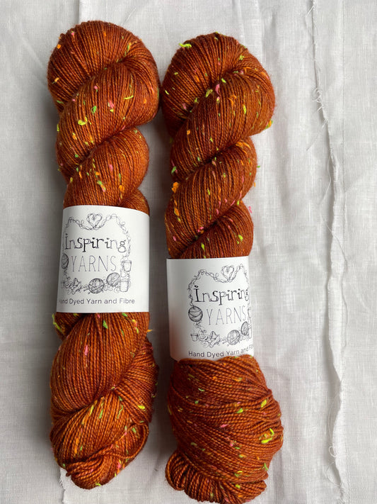 Two skeins of tonal burnt orange coloured hand dyed Disco Donegal yarn branded Inspiring Yarns on a white linen background.  The yarn has multi coloured neon neps.