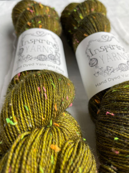 Close up of two skeins of tonal dark olive green colour  hand dyed Disco Donegal yarn branded Inspiring Yarns on a white linen background.  The yarn has multi coloured neon neps.