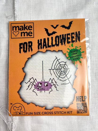 A mini crosstitch kit for a purple spider and a web photographed on a white linen background.  It is branded Mouseloft Make Me For Halloween in orange packaging.
