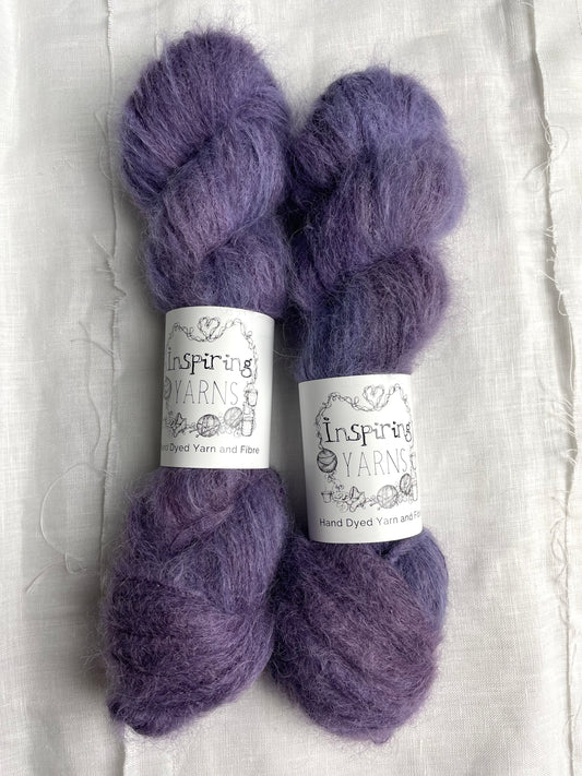 Two skeins of brushed suri silk yarn in a tonal dark purple colour labelled Inspiring Yarns on a white linen background. 