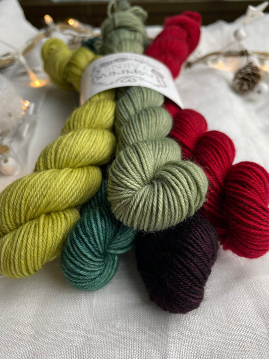 A set of five hand dyed yarn mini skeins on Merino Smooth Sock Yarn in red and green tones  labelled Inspiring Yarns.  The background is white linen and some Christmas lights with pinecones, twigs and stars.