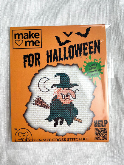 A mini crosstitch kit for a witch on a broomstick with a crescent moon photographed on a white linen background.  It is branded Mouseloft Make Me For Halloween in orange packaging.