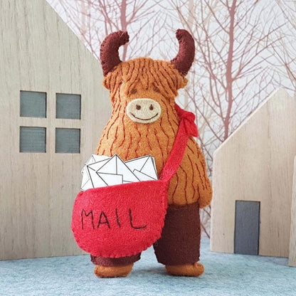 Stuffed highland cow with horns holding a red bag labeled 'MAIL' in front of wooden houses.