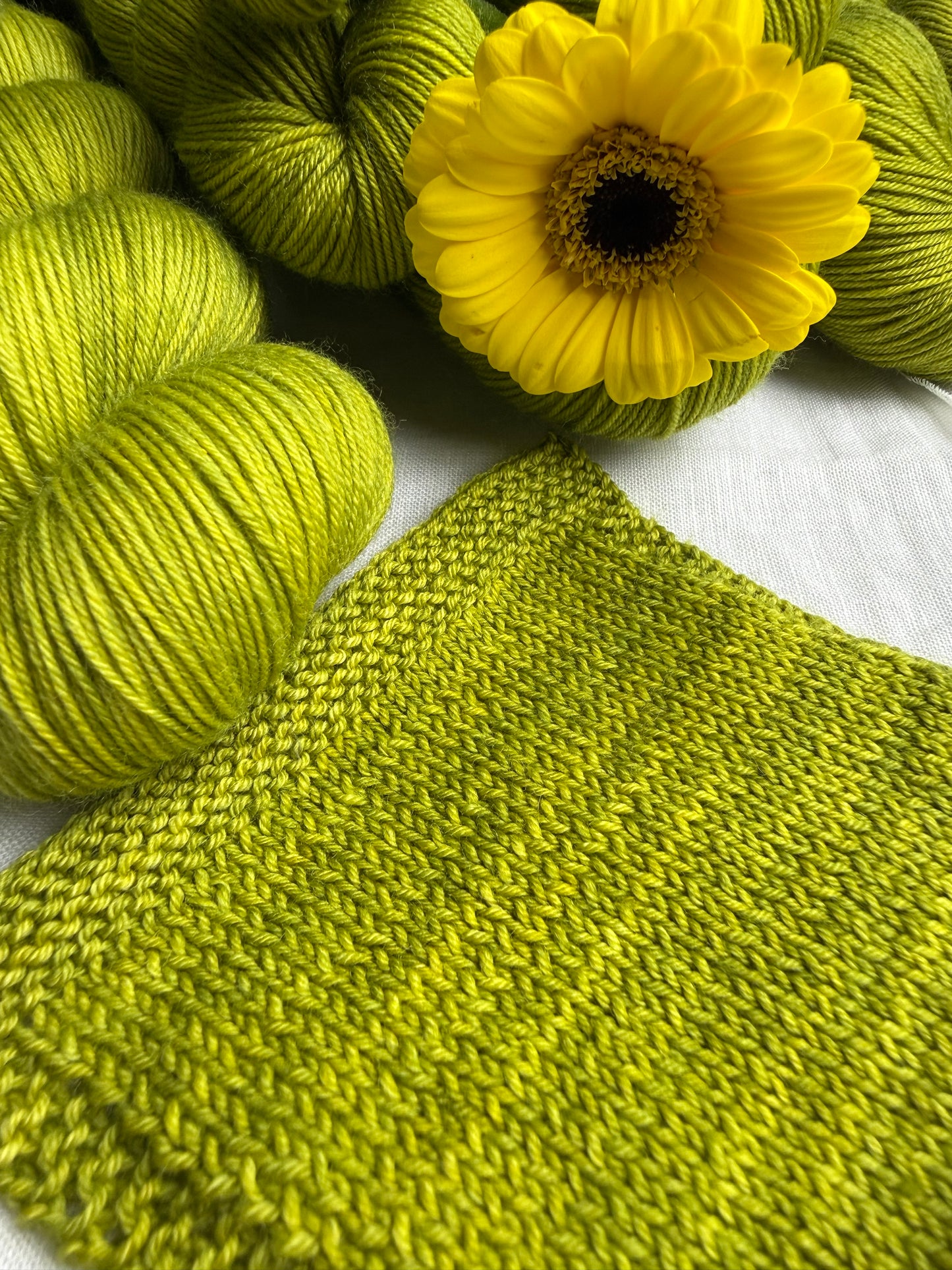 some skeins of bright green hand dyed yarn and a yellow flower with a knitted square swatch on a white linen background