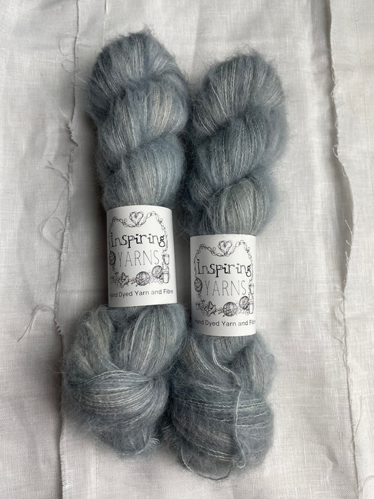 Two skeins of fluffy Brushed Suri Silk Lace yarn hand dyed in a pale blue grey colourway and labelled Inspiring Yarns on a white linen background.