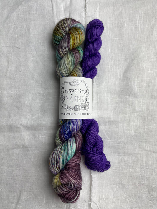 A set of hand dyed sock yarn comprising one 50g dark plum speckled and one 20g bright purple on a white linen background.  The yarn is branded Inspiring Yarns.