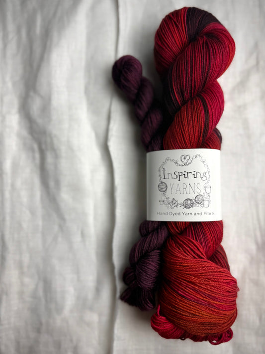 Variegated red, pink, orange and purple yarn skein with a burgundy mini skein labelled Inspiring Yarns on a white fabric background
