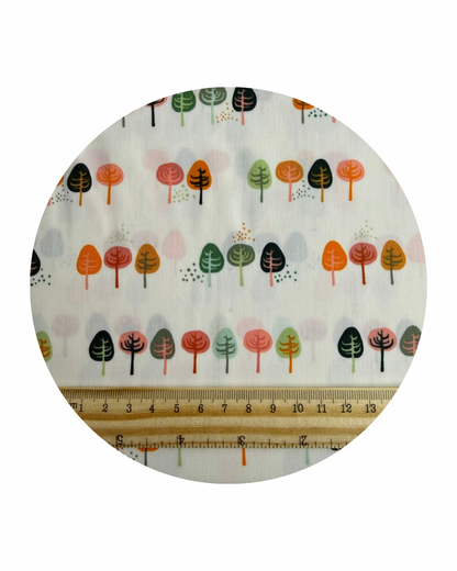 A circular swatch of Clear Path from the Lakeside Collection from Art Gallery Fabrics, a design of stylised small trees in rows in greens, oranges and pinks on a cream background and a wooden ruler in centimetres for scale on a white background.