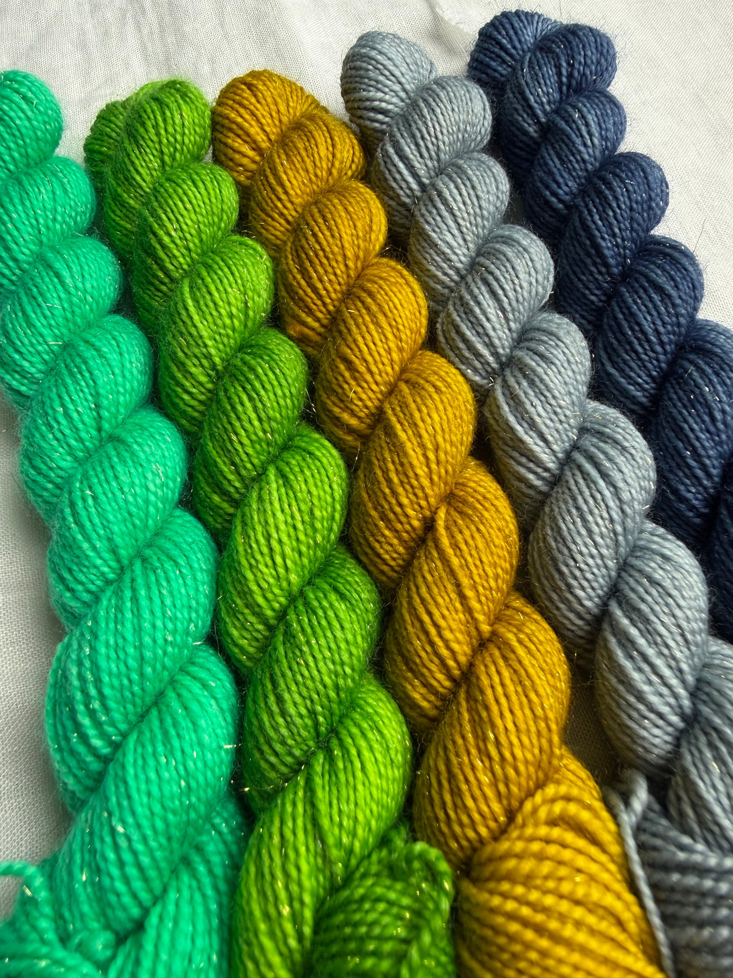 Five mini skeins of yarn in blue, blue grey, yellow, green & bright mint green on a light background