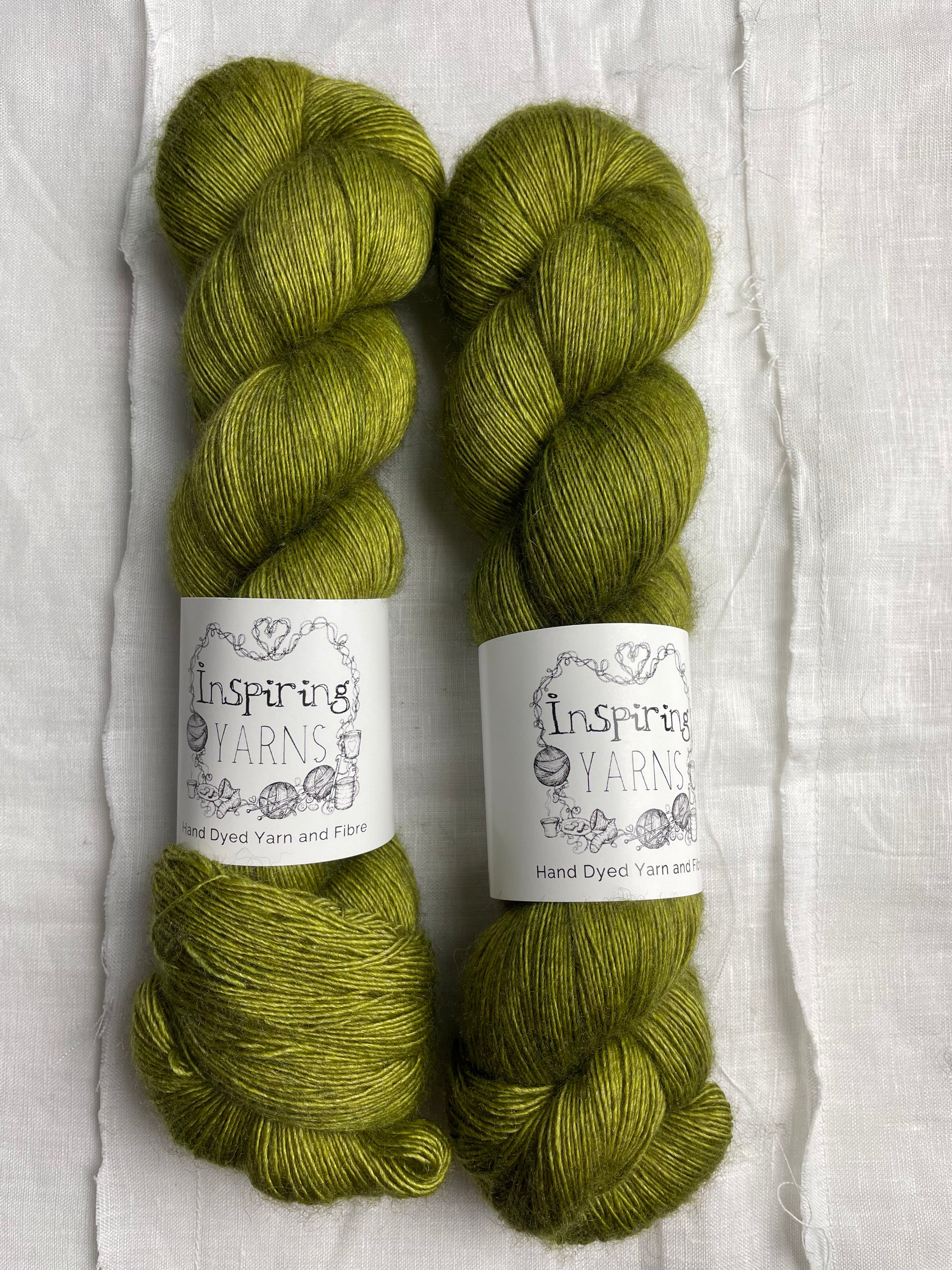 Two tonal spring green coloured skeins of hand dyed Yak Silk Singles branded Inspiring Yarns on a white linen background.