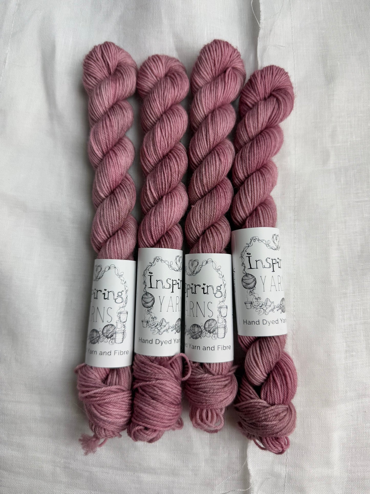 Four skeins of dusty pink yarn with inspiring yarn labels on a white fabric background.