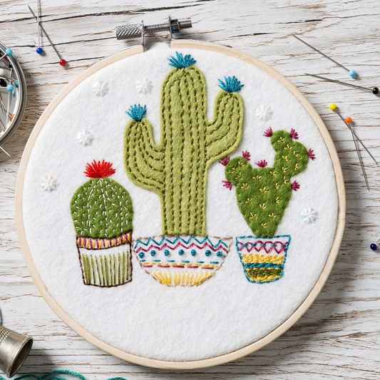 Embroidery hoop with a felt and embroidered design featuring 3 cactii in brightly coloured pots.  The hoop rests on a white wooden background with pins scattered around.