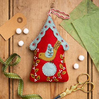 decorative felt bird house with embroidered details and a bird on the front.  on a wooden surface and a selection of crafting accessories.
