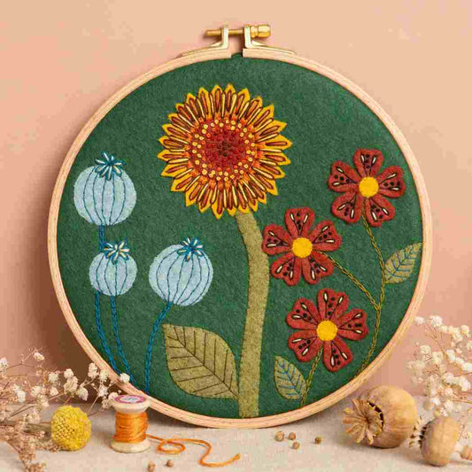 Embroidery hoop with a felt and embroidered design featuring botanical design on a dark green background.  The hoop rests on a terracotta background with a bunch of dried flowers and a spool of thread in the foreground.