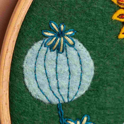 Embroidery of a light blue seed pod with a blue flower on a green fabric background
