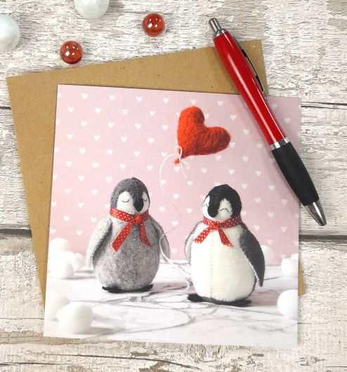 greeting card with an image of 2 felt penguins with a heart balloon on a wooden background with a pen and envelope.