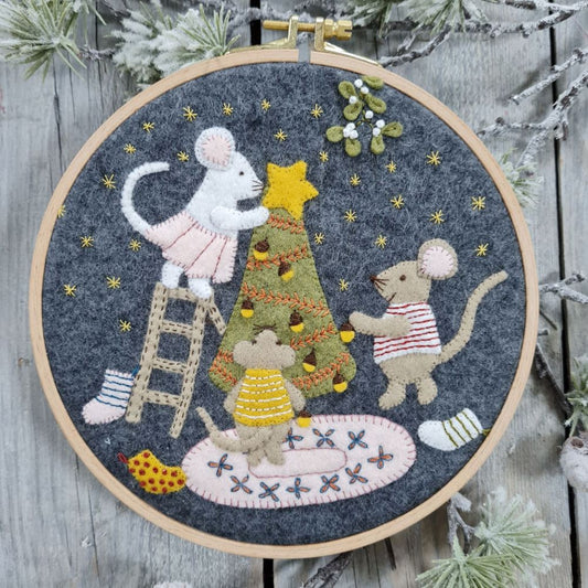 Embroidery hoop with a felt and embroidered Christmas design featuring 3 mice decorating a tree.  The hoop rests on a white wooden background with some fir tree branches.