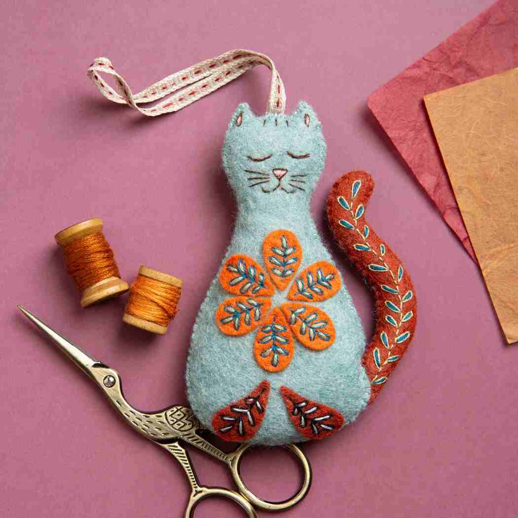 decorative felt cat with embroidered details on a plain pink background surrounded by a selection of sewing accessories.