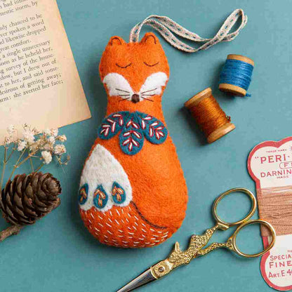 decorative felt fox decoration with embroidered details on a plain blue background surrounded by a selection of sewing accessories.