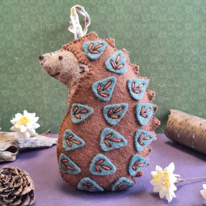 Felted hedgehog ornament with embroidered details on a purple surface with flowers and pinecones.