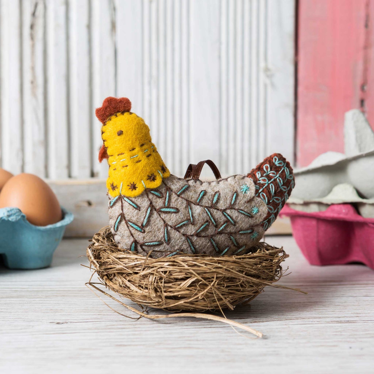 decorative felt rooster in a nest with eggs in the background on a white and red surface 
