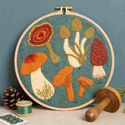 Embroidery hoop with a felt and embroidered design featuring autumnal colour fungi on a blue background.  The hoop rests on a blue background with some sewing accessories in the foreground.