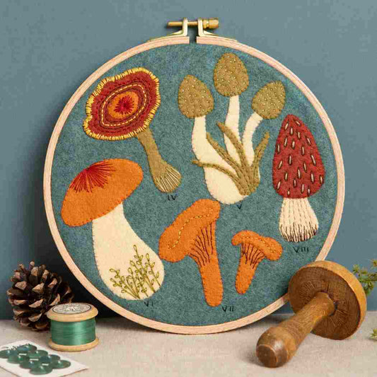 Embroidery hoop with a felt and embroidered design featuring autumnal colour fungi on a blue background.  The hoop rests on a blue background with some sewing accessories in the foreground.