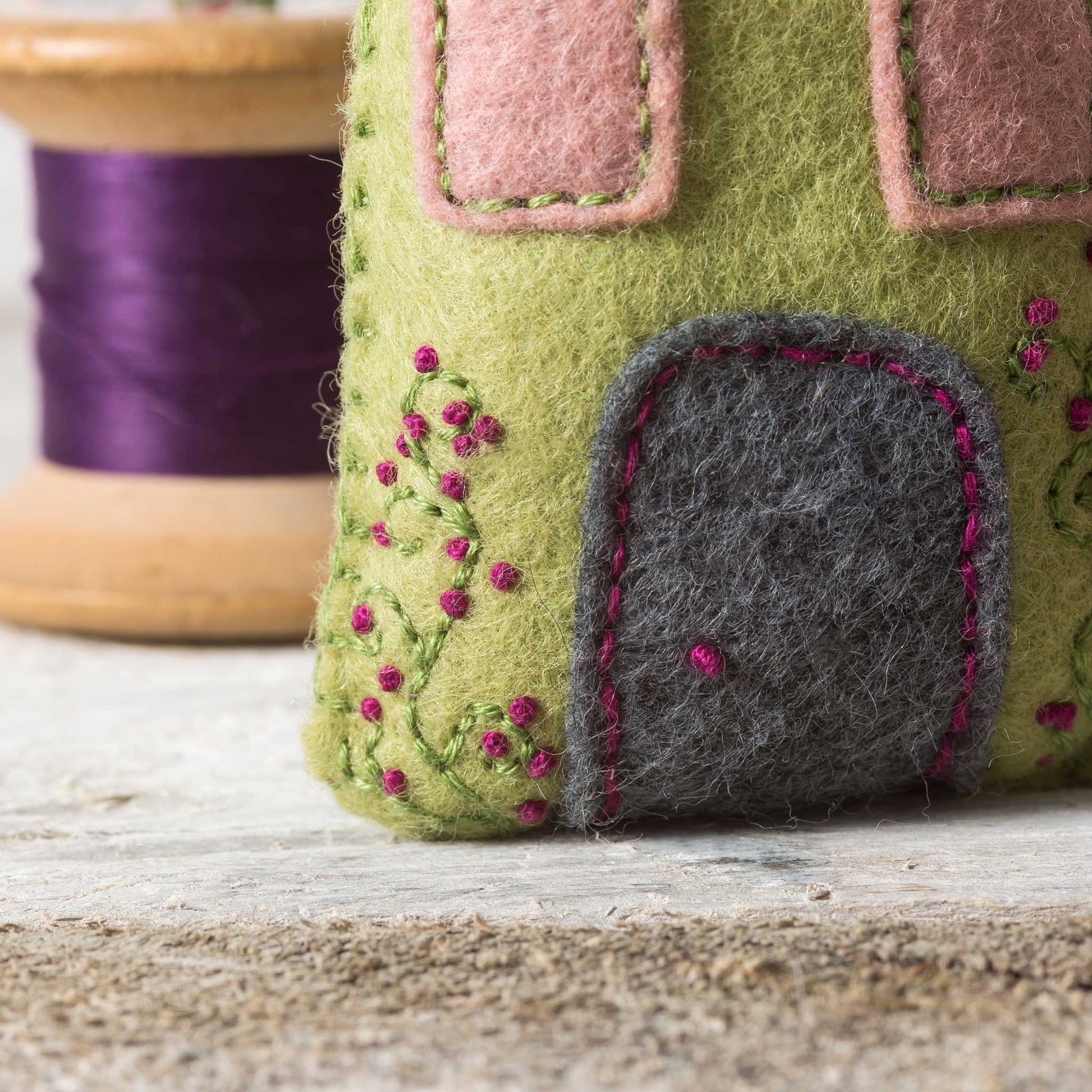 Felted green house with pink windows and gray door on a wooden surface with purple thread spool in the background.