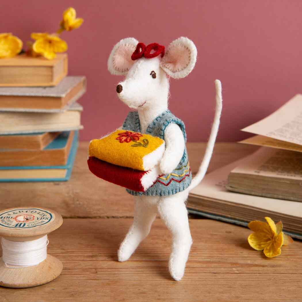 Stuffed mouse toy with a bow and small bag on a wooden surface with books and flowers.  