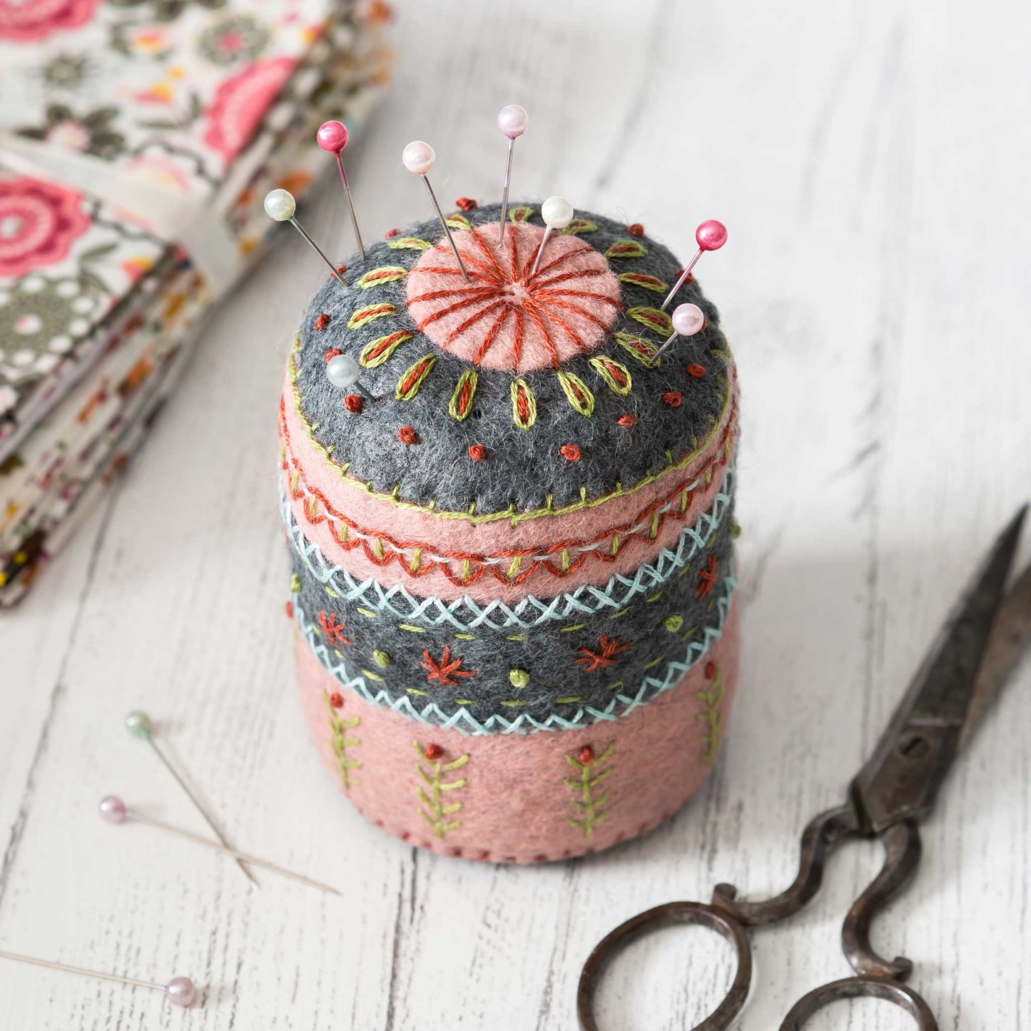pincushion on white wooden surface with stack of fabric and scissors in the corners.