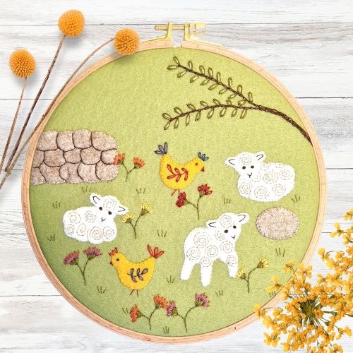 Embroidery hoop with a felt and embroidered design featuring sheep and chickens in a meadow.  The hoop rests on a white wooden background with a bunch of yellow flowers.
