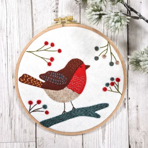 Embroidery hoop with a felt and embroidered design featuring a robin on a branch and some berries.  The hoop rests on a white wooden background with a branch of snowy fir tree..