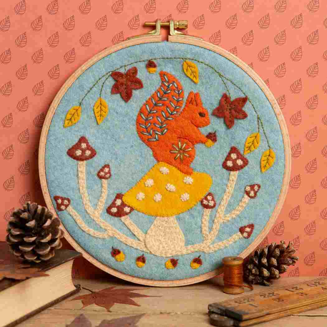 Embroidery hoop with a felt and embroidered design featuring a squirrel on some toadstools in autumnal colours.  The hoop rests on a terracotta leaf print background with pinecones, books, thread  and a wooden ruler in the foreground. 