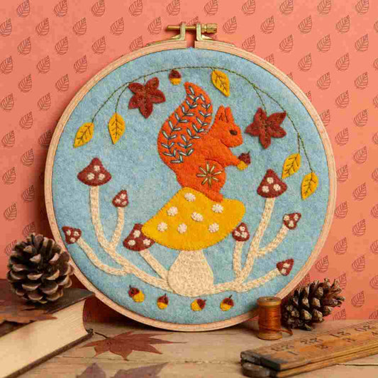 Embroidery hoop with a felt and embroidered design featuring a squirrel on some toadstools in autumnal colours.  The hoop rests on a terracotta leaf print background with pinecones, books, thread  and a wooden ruler in the foreground. 